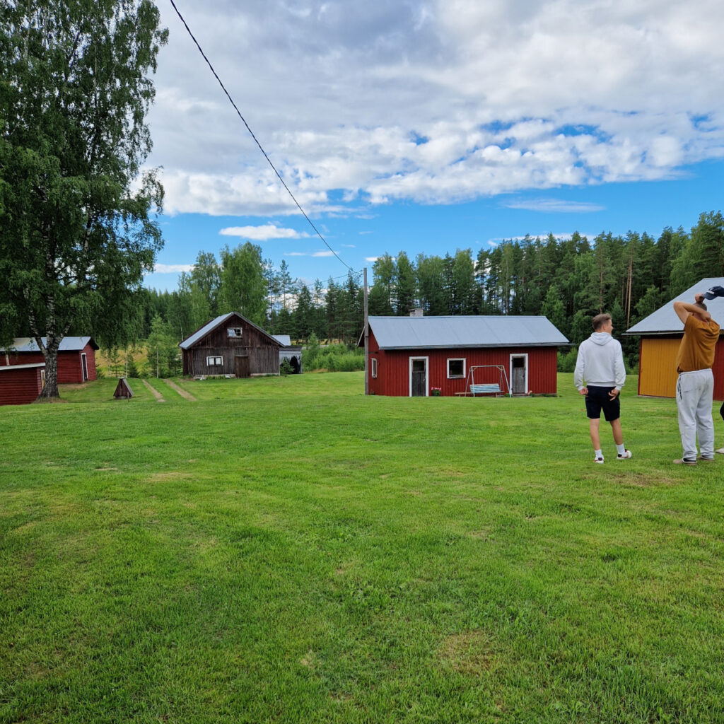 Mökkimaisema, jossa kaksi miestä seisoo nurmella ja taustalla on mökkejä
